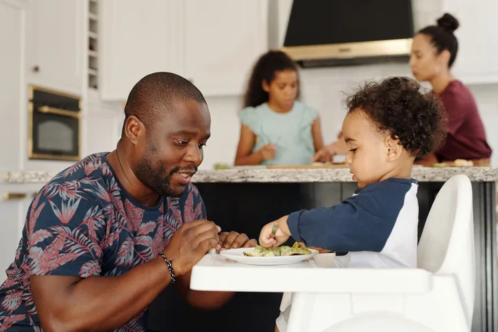 A parent sitting beside a toddler in a high chair at a family mealtime at home