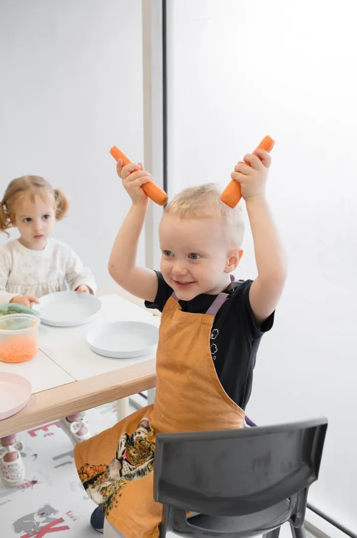 A smiling toddler holding up two carrot sticks at the mealtime table