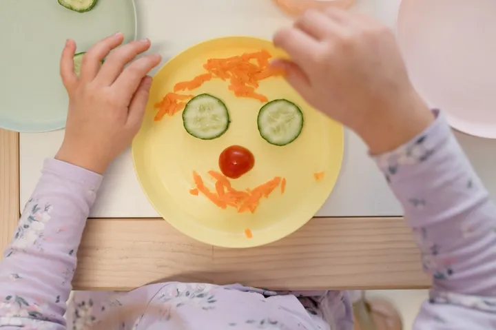 A child arranging grated carrot, cucumber and tomato into a face on a yellow plate