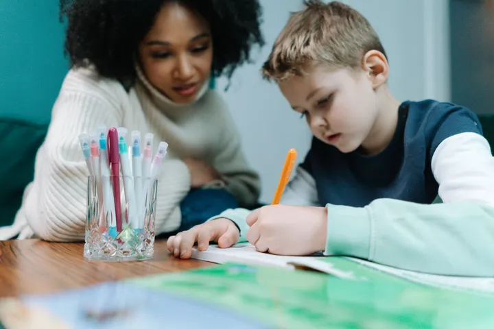 A speech pathologist sitting beside a young boy while he writes at a table at home
